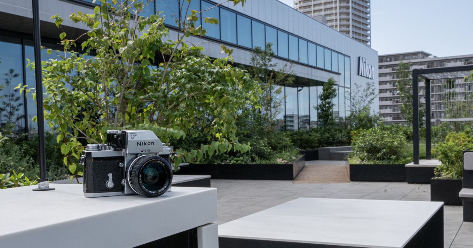 A Nikon camera sits on a white table outdoors with a modern building and greenery in the background. The building has large glass windows and a visible Nikon logo. A Nikon camera sits on a white table outdoors with a modern building and greenery in the background. The building has large glass windows and a visible Nikon logo.