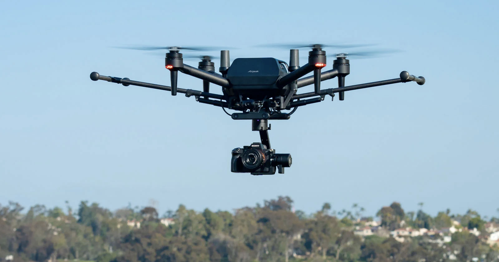 A quadcopter drone equipped with a camera hovers in the sky. The background shows a blurred landscape of trees and buildings under a clear blue sky. A quadcopter drone equipped with a camera hovers in the sky. The background shows a blurred landscape of trees and buildings under a clear blue sky.