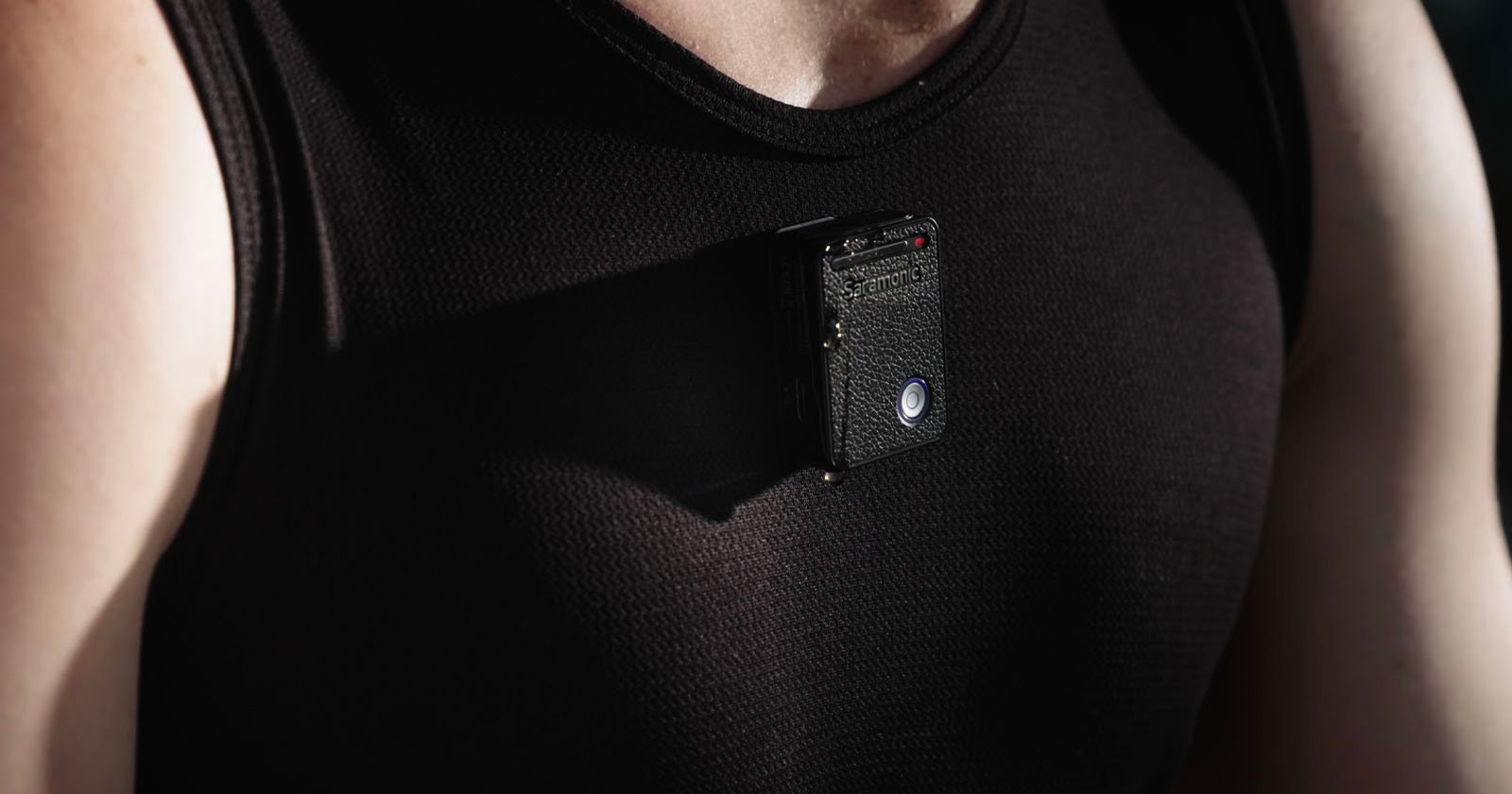 A person wearing a black tank top with a small camera device attached to the chest area, capturing an image in a dark, shaded setting. A person wearing a black tank top with a small camera device attached to the chest area, capturing an image in a dark, shaded setting.