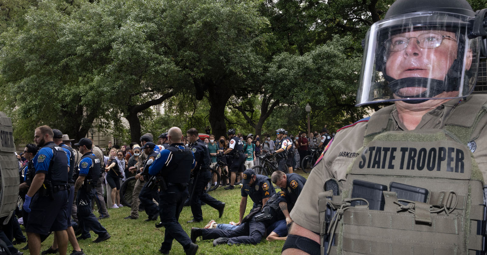 A state trooper in riot gear stands in the foreground, while a group of police officers and a crowd of protesters are in the background on a grassy area. Trees line the scene, creating a natural backdrop. A state trooper in riot gear stands in the foreground, while a group of police officers and a crowd of protesters are in the background on a grassy area. Trees line the scene, creating a natural backdrop.