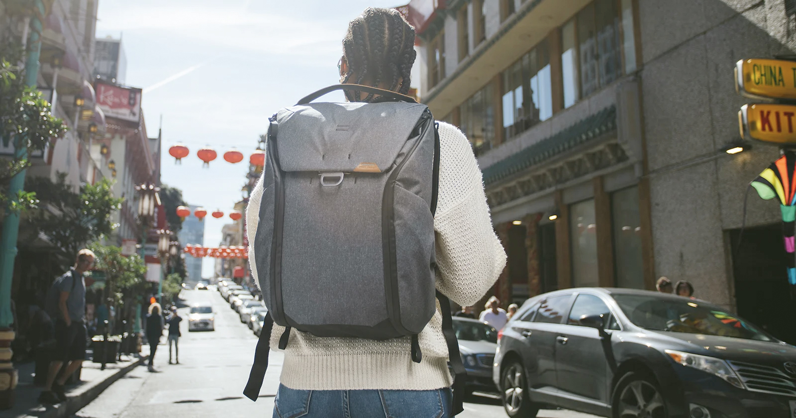 Person with a gray backpack walking down a busy city street lined with vehicles and pedestrians. The street has red lanterns hanging above, and signs in Chinese characters are visible on buildings. The person wears a white sweater and jeans. Person with a gray backpack walking down a busy city street lined with vehicles and pedestrians. The street has red lanterns hanging above, and signs in Chinese characters are visible on buildings. The person wears a white sweater and jeans.