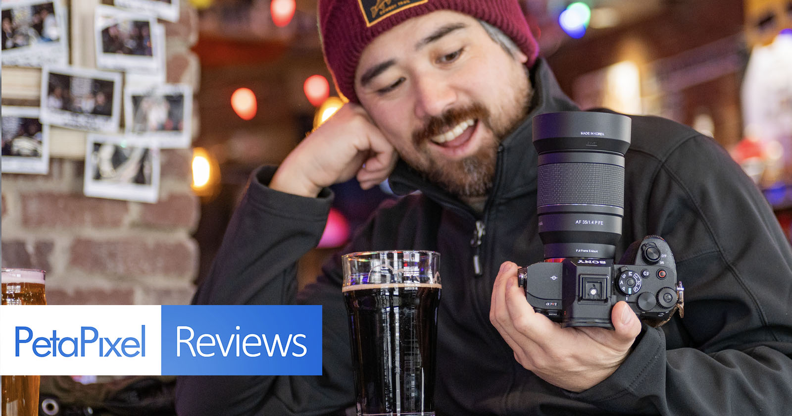 A smiling person in a beanie and black jacket holds a camera in one hand, while sitting at a table with a glass of dark beer. The background features a brick wall with photos and warm lighting. Text: PetaPixel Reviews. A smiling person in a beanie and black jacket holds a camera in one hand, while sitting at a table with a glass of dark beer. The background features a brick wall with photos and warm lighting. Text: PetaPixel Reviews.