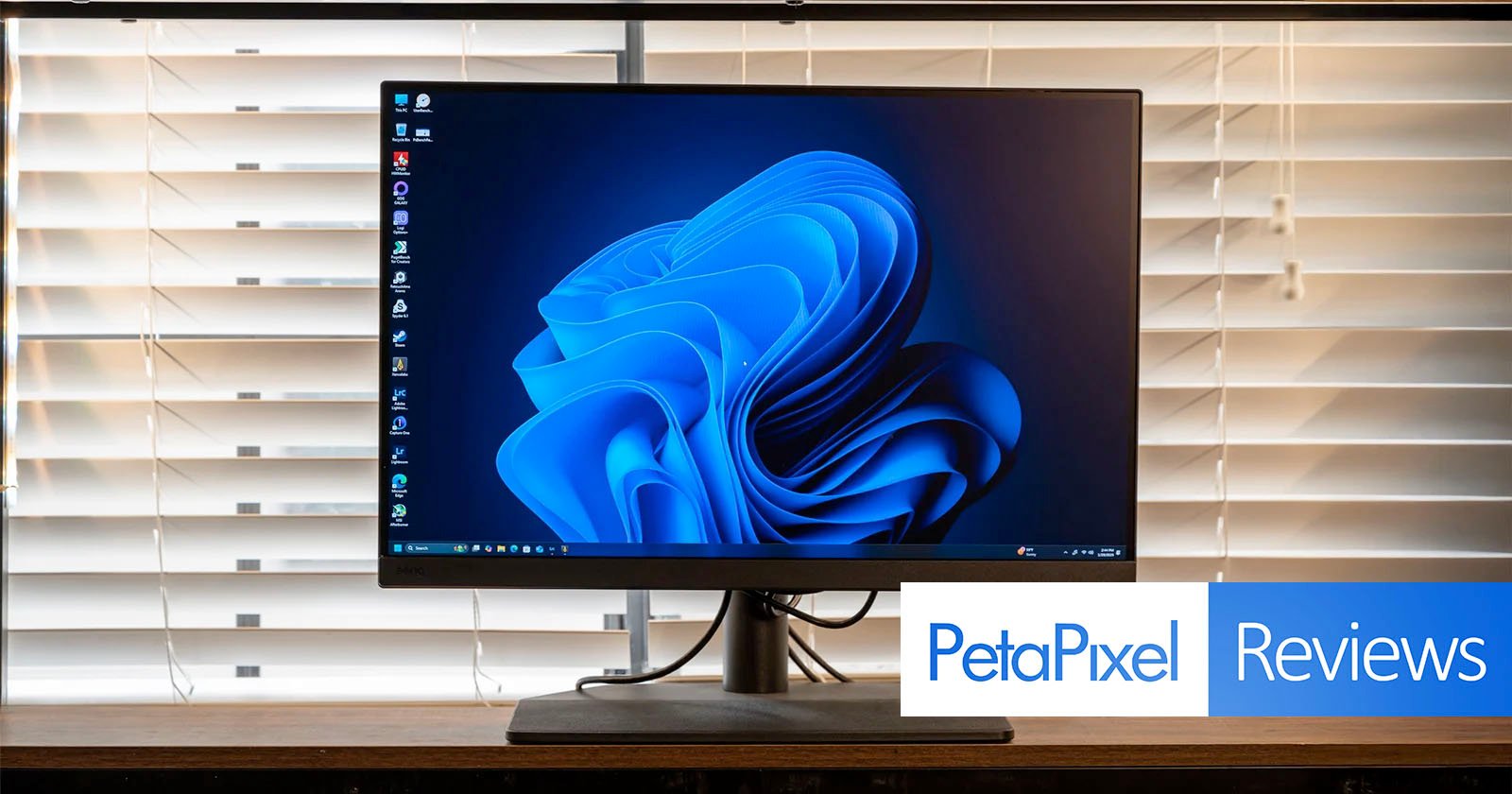 A desktop computer monitor displaying a swirling blue abstract design on its screen. The monitor is placed on a wooden table in front of white blinds. The image has the text PetaPixel Reviews overlaid in the bottom right corner. A desktop computer monitor displaying a swirling blue abstract design on its screen. The monitor is placed on a wooden table in front of white blinds. The image has the text PetaPixel Reviews overlaid in the bottom right corner.