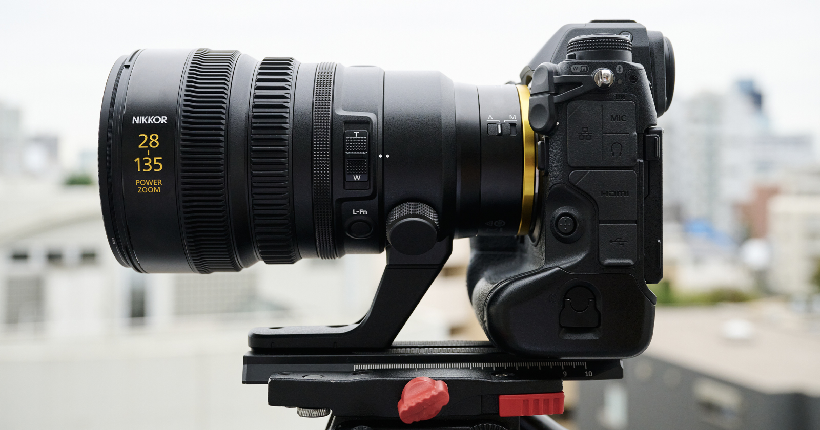 Side view of a professional camera with a large zoom lens mounted on a tripod. The lens has 28 135 and Power Zoom marked on it. The background is a blurred urban landscape, suggesting an outdoor setting. Side view of a professional camera with a large zoom lens mounted on a tripod. The lens has 28 135 and Power Zoom marked on it. The background is a blurred urban landscape, suggesting an outdoor setting.