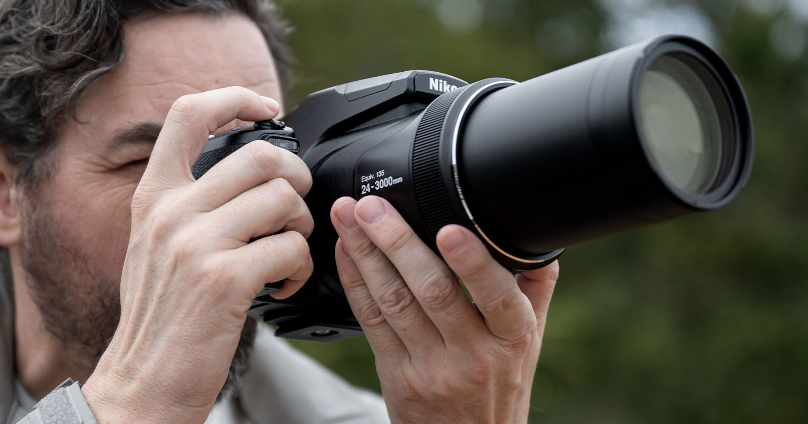A person is holding a Nikon camera with a large lens, capturing a photo. The individual appears focused, with the lens pointed forward. The background is softly blurred with greenery. A person is holding a Nikon camera with a large lens, capturing a photo. The individual appears focused, with the lens pointed forward. The background is softly blurred with greenery.