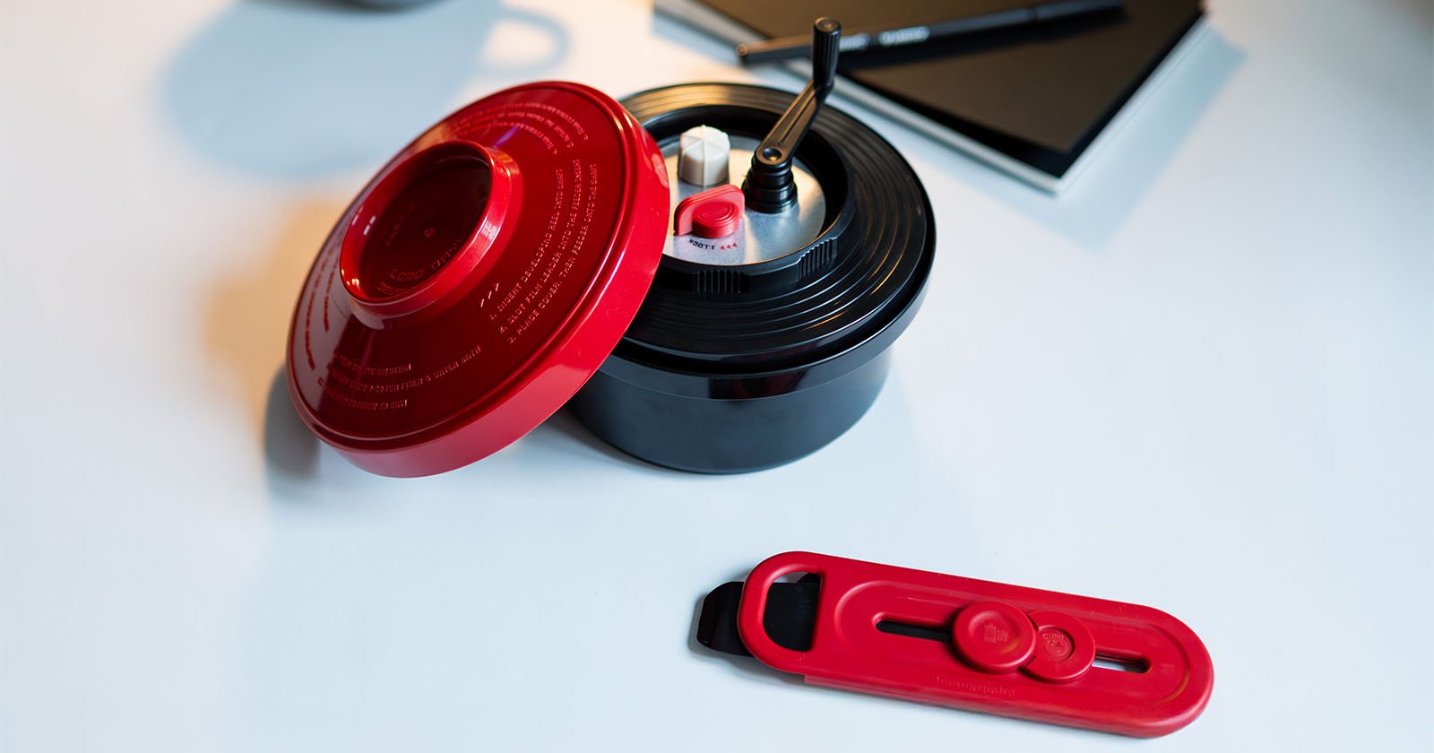 A red and black kitchen gadget with a lid and crank handle is on a light surface. A matching red attachment lies nearby. A notebook and pen are in the background. A red and black kitchen gadget with a lid and crank handle is on a light surface. A matching red attachment lies nearby. A notebook and pen are in the background.