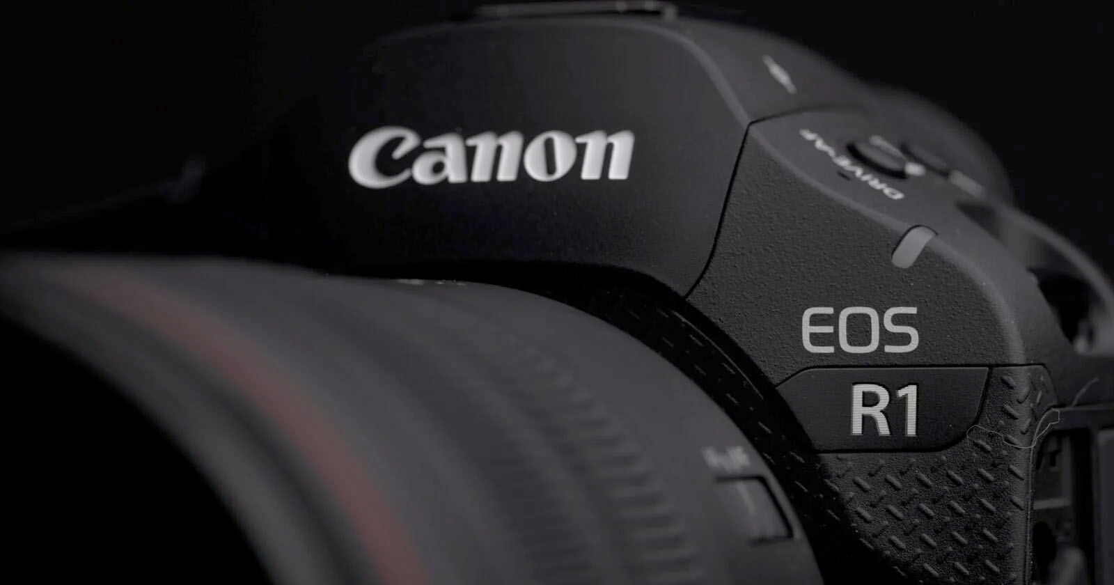 Close-up of a Canon EOS R1 camera, showcasing the brand logo and model name on a textured black body, with part of the attached lens visible on the left. Close-up of a Canon EOS R1 camera, showcasing the brand logo and model name on a textured black body, with part of the attached lens visible on the left.