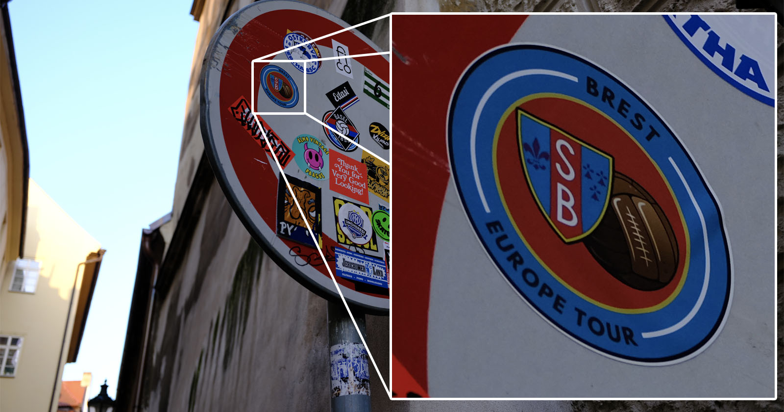 Close-up of a street sign covered with various colorful stickers. The sign is red with a white circle, partially visible, mounted on a metal pole. Stickers include a logo with overlapping letters SB and text Brest Europe Tour. Close-up of a street sign covered with various colorful stickers. The sign is red with a white circle, partially visible, mounted on a metal pole. Stickers include a logo with overlapping letters SB and text Brest Europe Tour.