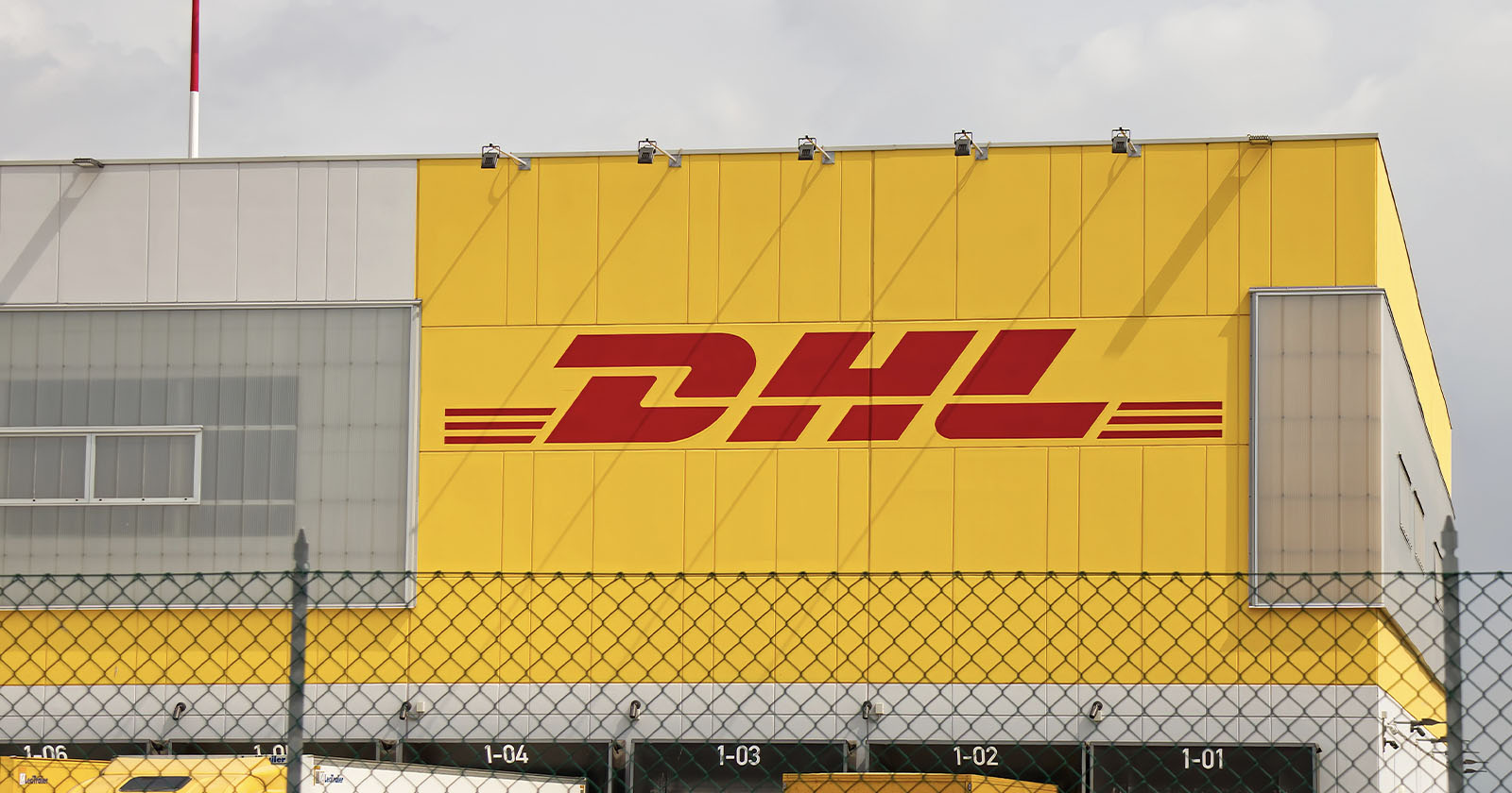 A large yellow DHL building with the company’s red logo on the side, viewed behind a chain-link fence under a cloudy sky. A large yellow DHL building with the company’s red logo on the side, viewed behind a chain-link fence under a cloudy sky.