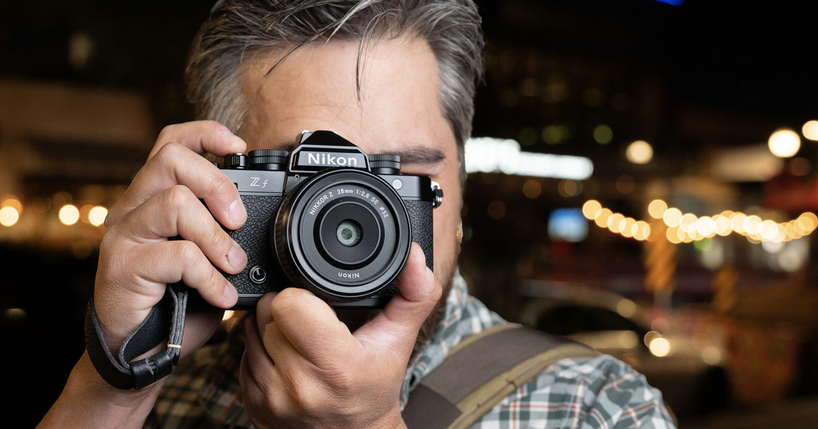 A person with dark hair and a beard holds a Nikon Zfc camera up to their face, ready to take a photo. The background is blurred with warm, glowing lights. A person with dark hair and a beard holds a Nikon Zfc camera up to their face, ready to take a photo. The background is blurred with warm, glowing lights.