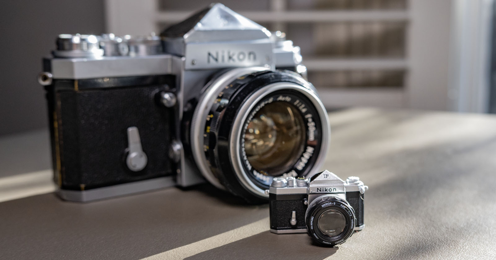 A large Nikon camera with a classic design is placed beside a miniature version on a table. The room in the background is softly lit, highlighting the intricate details of both cameras. A large Nikon camera with a classic design is placed beside a miniature version on a table. The room in the background is softly lit, highlighting the intricate details of both cameras.