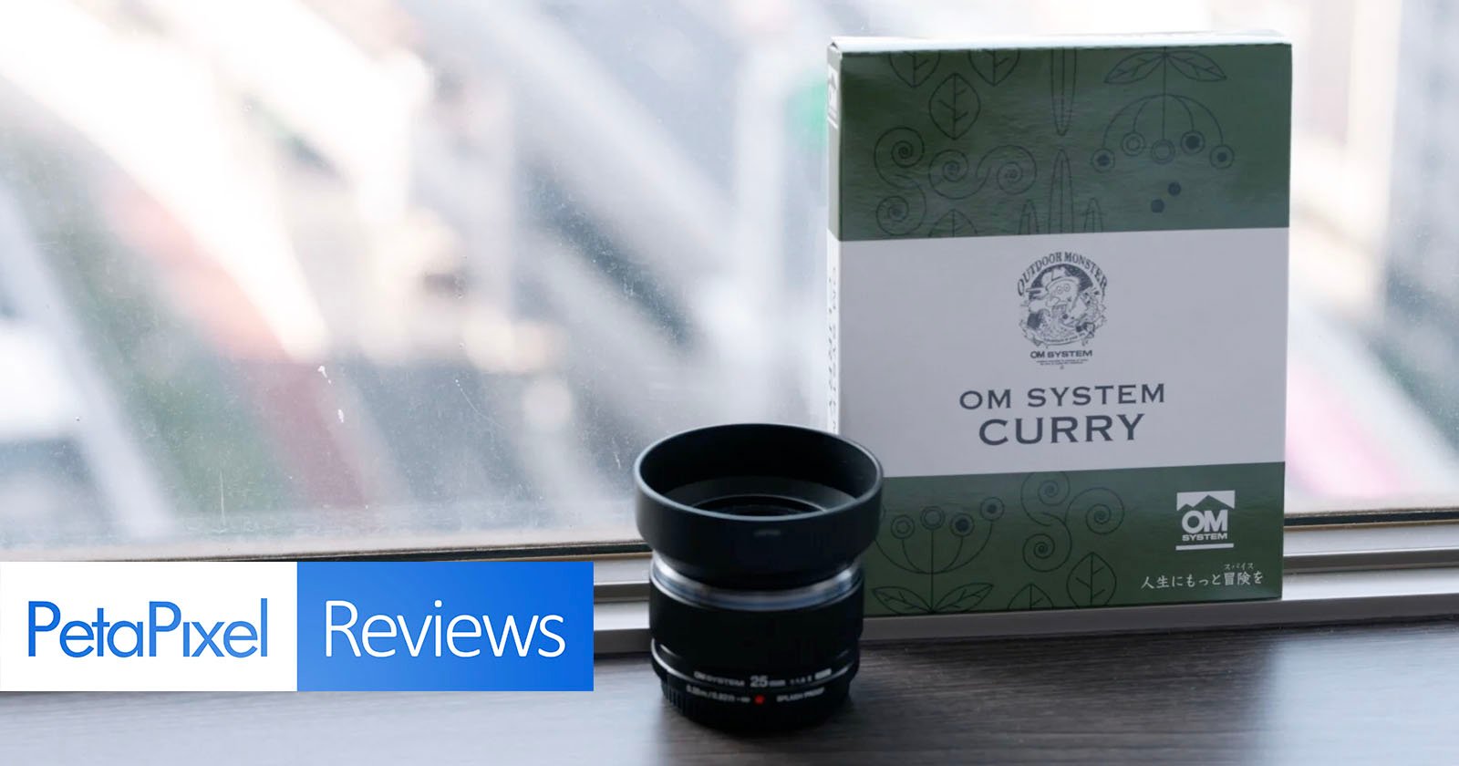 A camera lens and a box labeled OM System Curry are placed on a windowsill. The background shows a blurred cityscape, while the foreground features a PetaPixel Reviews banner. A camera lens and a box labeled OM System Curry are placed on a windowsill. The background shows a blurred cityscape, while the foreground features a PetaPixel Reviews banner.