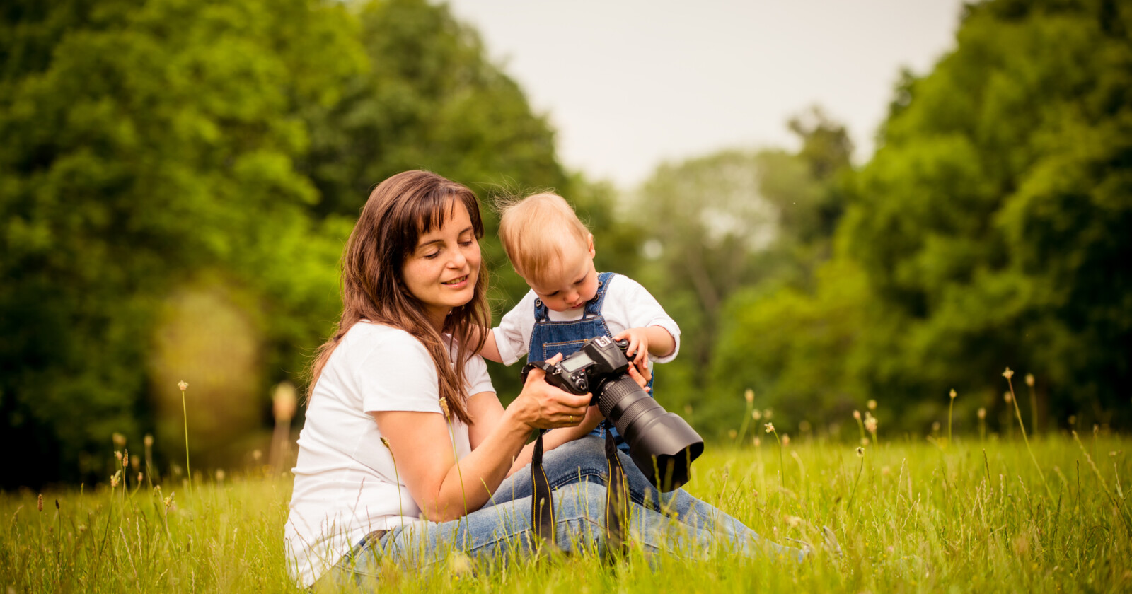 A woman and a young child sit on grass in a park, looking at a camera together. The woman smiles while the child touches the camera, surrounded by green trees and a peaceful, natural setting. A woman and a young child sit on grass in a park, looking at a camera together. The woman smiles while the child touches the camera, surrounded by green trees and a peaceful, natural setting.