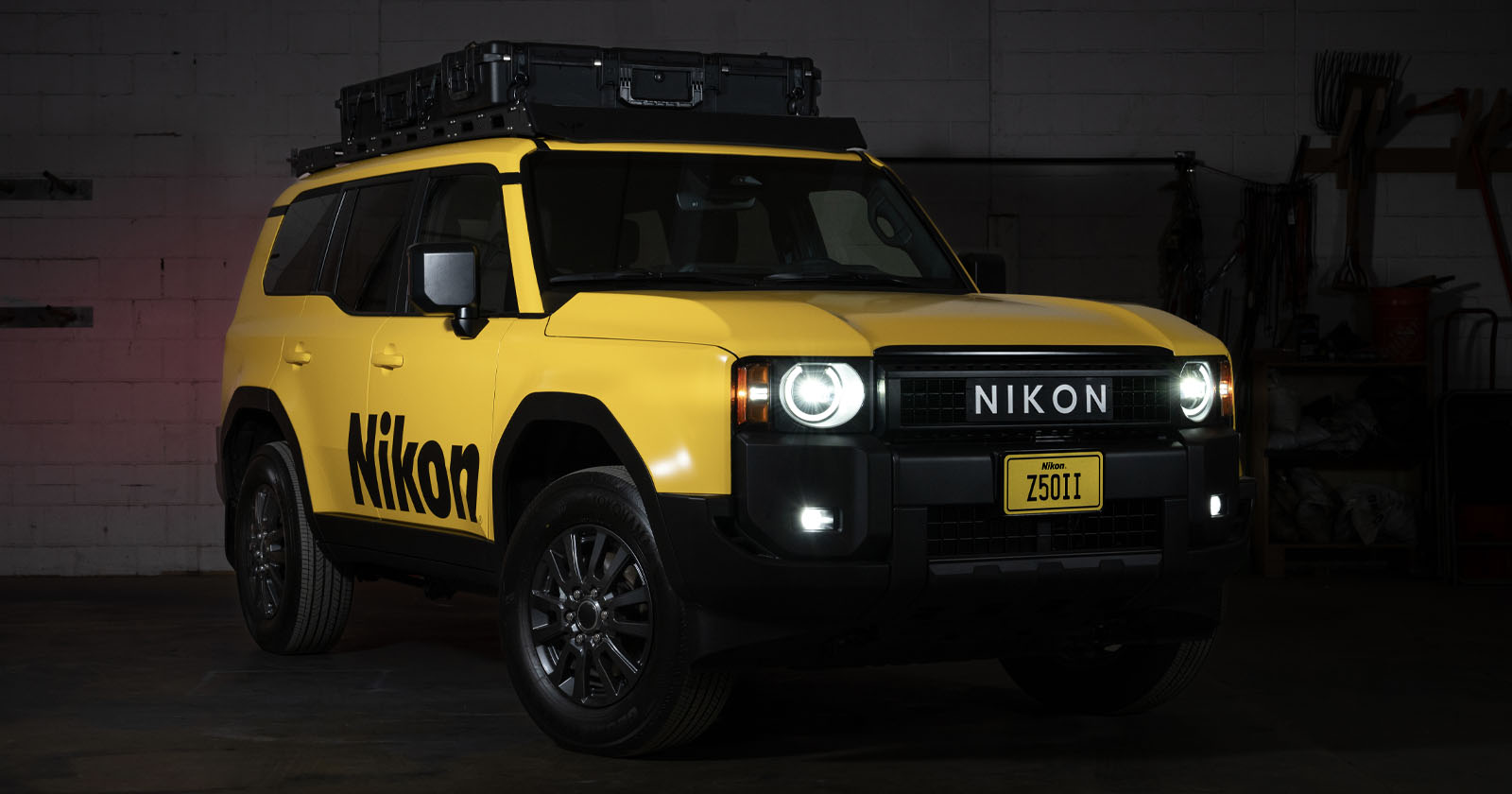 A yellow SUV with “Nikon” branding on the side and front, parked indoors. The vehicle has a roof storage box, black wheels, and a custom license plate reading “Z50II.” The headlights and fog lights are on. A yellow SUV with “Nikon” branding on the side and front, parked indoors. The vehicle has a roof storage box, black wheels, and a custom license plate reading “Z50II.” The headlights and fog lights are on.
