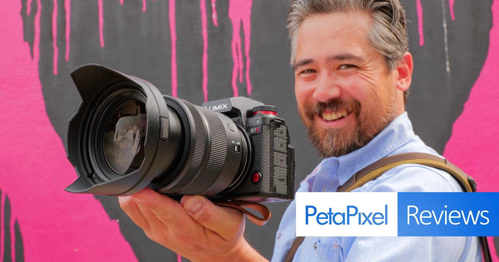 A smiling man with a beard holds a large Lumix camera towards the viewer, standing in front of a pink and black painted wall. The image features the PetaPixel Reviews logo in the lower right corner. A smiling man with a beard holds a large Lumix camera towards the viewer, standing in front of a pink and black painted wall. The image features the PetaPixel Reviews logo in the lower right corner.