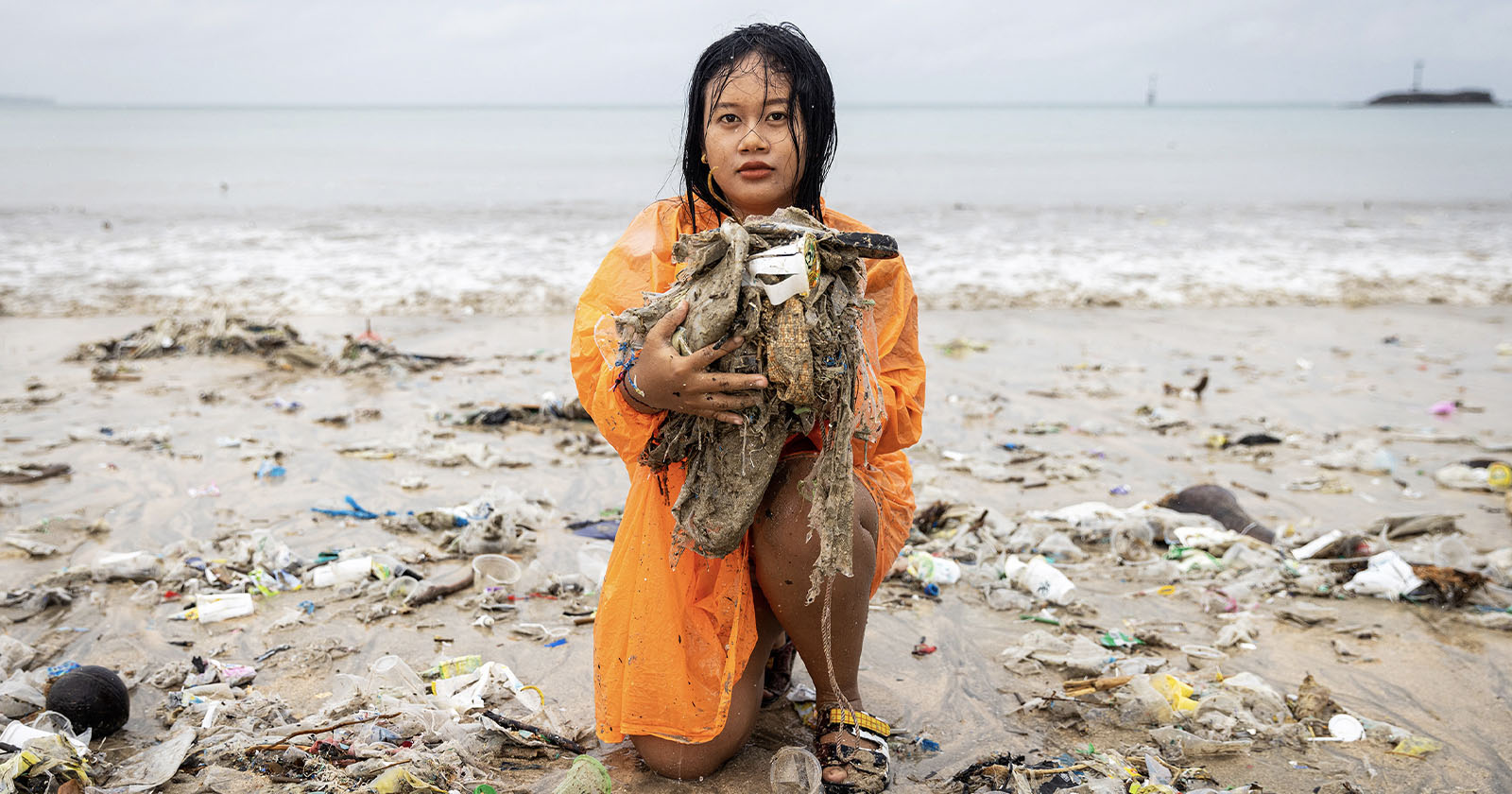 A woman in an orange raincoat kneels on a beach holding a large pile of trash, surrounded by litter and plastic debris, with the ocean and cloudy sky in the background. A woman in an orange raincoat kneels on a beach holding a large pile of trash, surrounded by litter and plastic debris, with the ocean and cloudy sky in the background.