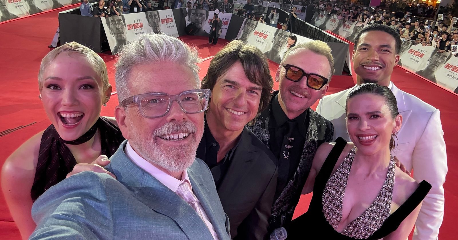 A group of six people in formal attire take a selfie together on a red carpet, with a crowd and event banners visible in the background. A group of six people in formal attire take a selfie together on a red carpet, with a crowd and event banners visible in the background.