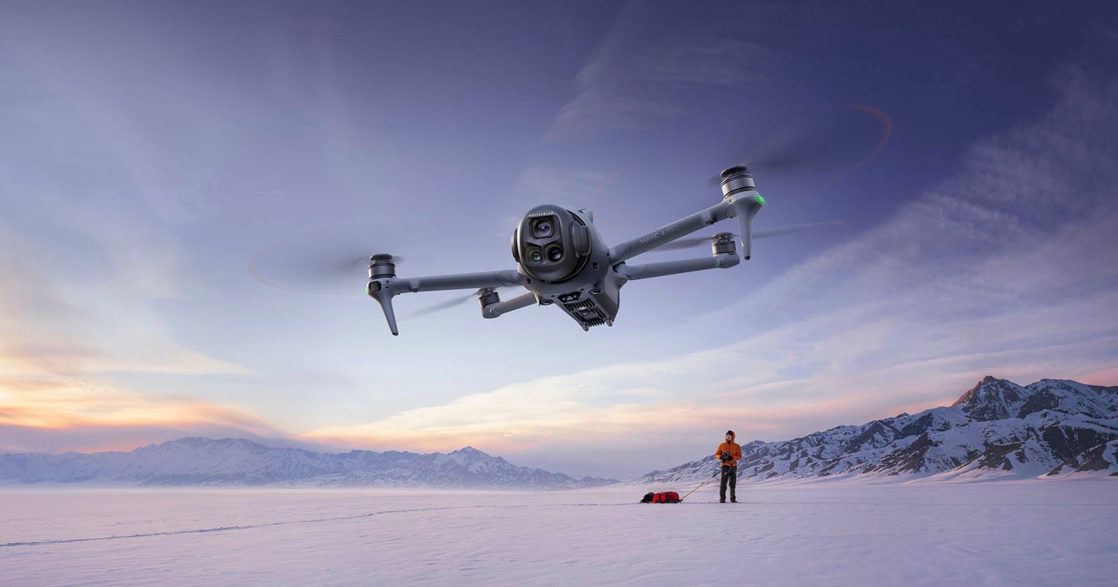 A drone hovers in the foreground above a snowy landscape at sunset, while a person stands on the snow below near equipment with mountains visible in the background. A drone hovers in the foreground above a snowy landscape at sunset, while a person stands on the snow below near equipment with mountains visible in the background.