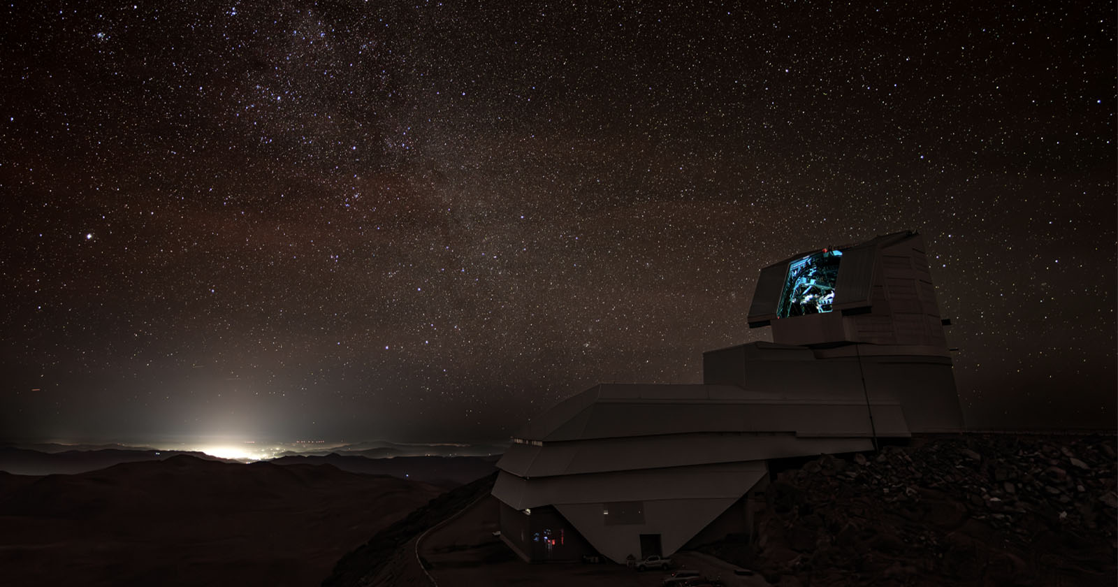 A large astronomical observatory sits atop a mountain under a star-filled night sky. In the distance, city lights glow, while the observatory’s dome is partially open, illuminated with a faint blue light inside. A large astronomical observatory sits atop a mountain under a star-filled night sky. In the distance, city lights glow, while the observatory’s dome is partially open, illuminated with a faint blue light inside.