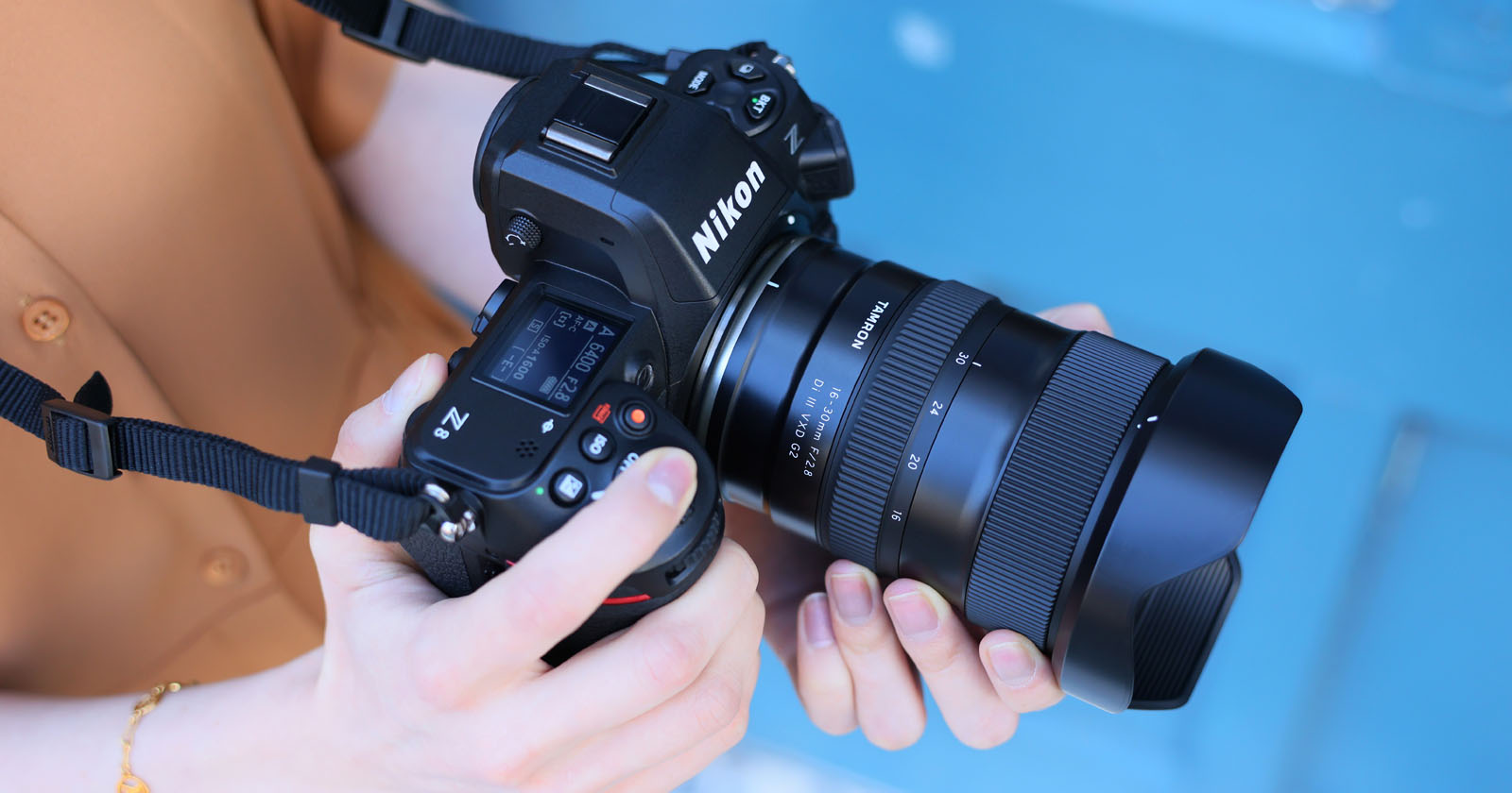 A person holds a Nikon Z series camera with a Sigma lens attached, against a blurred blue background. The camera is positioned for shooting, and the person's hands are visible adjusting the controls. A person holds a Nikon Z series camera with a Sigma lens attached, against a blurred blue background. The camera is positioned for shooting, and the person's hands are visible adjusting the controls.
