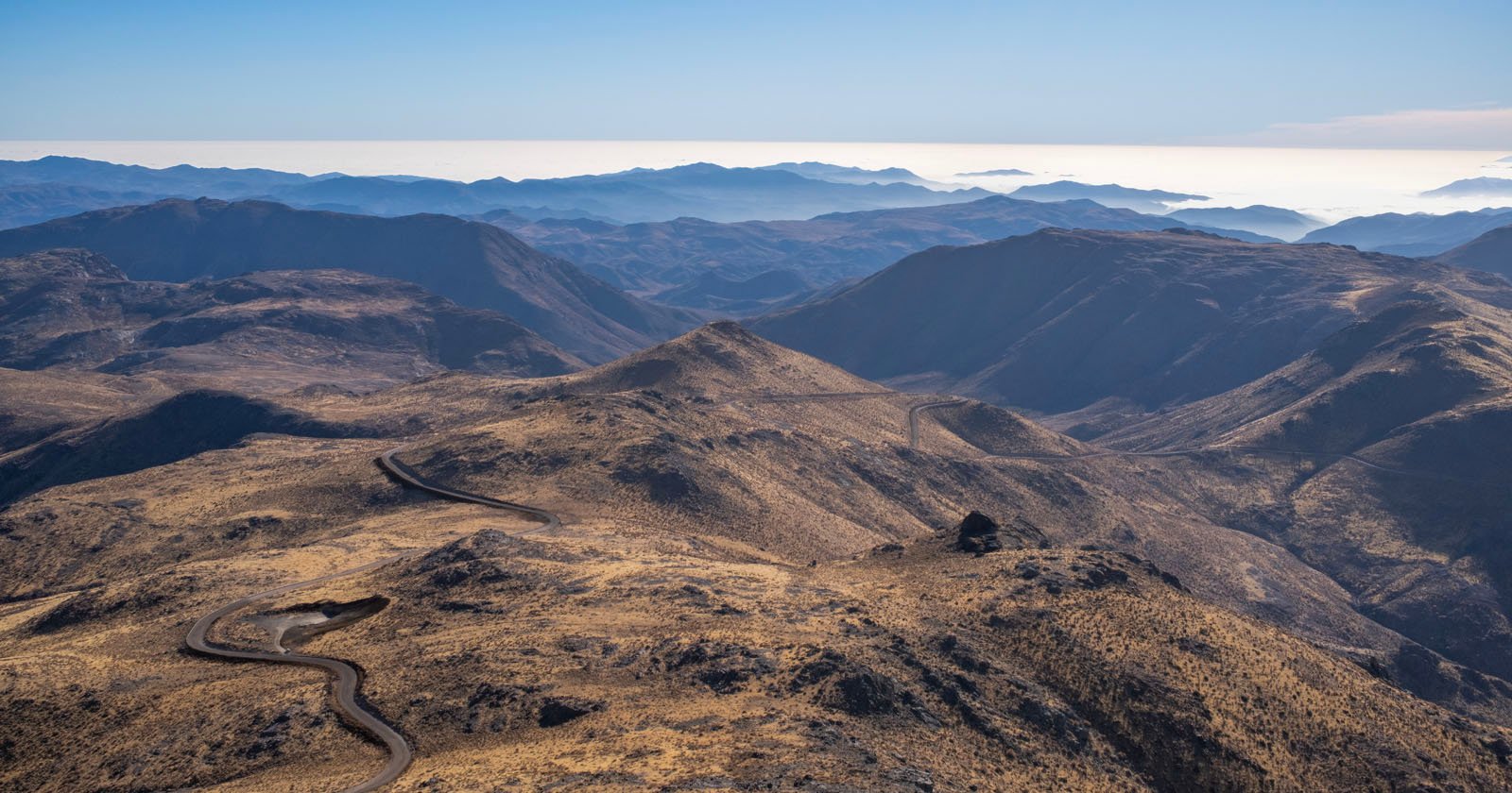A winding road cuts through dry, rugged mountains under a clear blue sky, with layers of distant hills and mist visible on the horizon. A winding road cuts through dry, rugged mountains under a clear blue sky, with layers of distant hills and mist visible on the horizon.