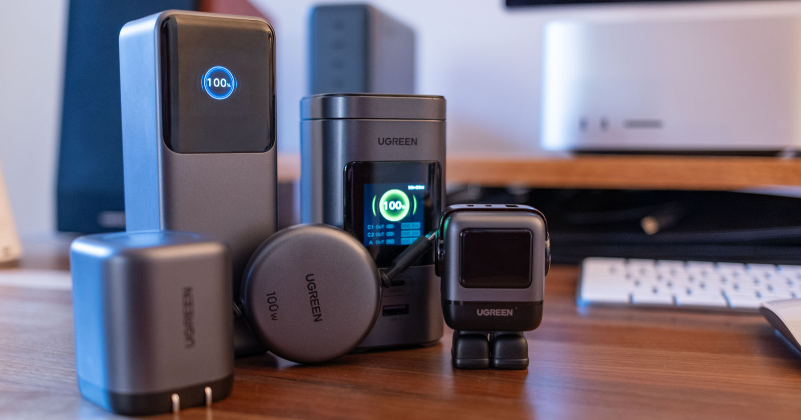 Several UGREEN electronic devices, including chargers and adapters, are displayed on a wooden desk in front of a computer setup, with digital screens showing charging icons. Several UGREEN electronic devices, including chargers and adapters, are displayed on a wooden desk in front of a computer setup, with digital screens showing charging icons.