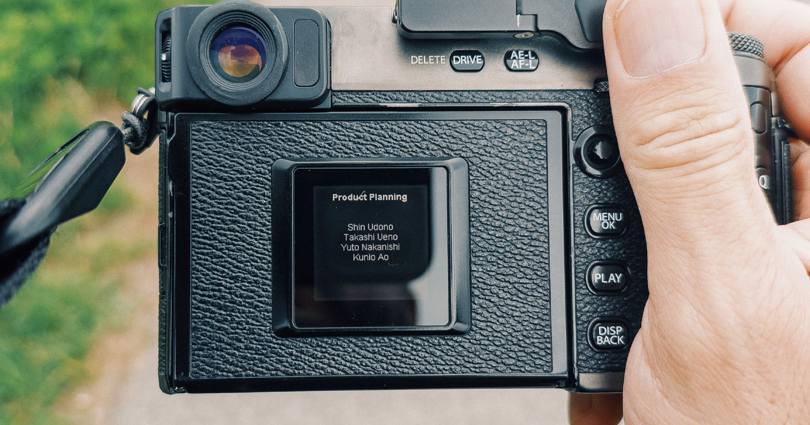 A close-up of a person holding a digital camera, showing its screen displaying a “Product Planning” credits list with four names. The camera’s buttons and textured grip are visible, with a blurred outdoor background. A close-up of a person holding a digital camera, showing its screen displaying a “Product Planning” credits list with four names. The camera’s buttons and textured grip are visible, with a blurred outdoor background.