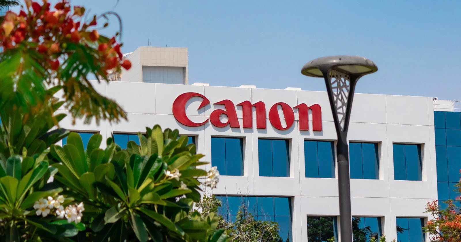 The exterior of a modern white building with large windows and a red Canon logo, surrounded by green trees and flowers, under a clear blue sky. The exterior of a modern white building with large windows and a red Canon logo, surrounded by green trees and flowers, under a clear blue sky.