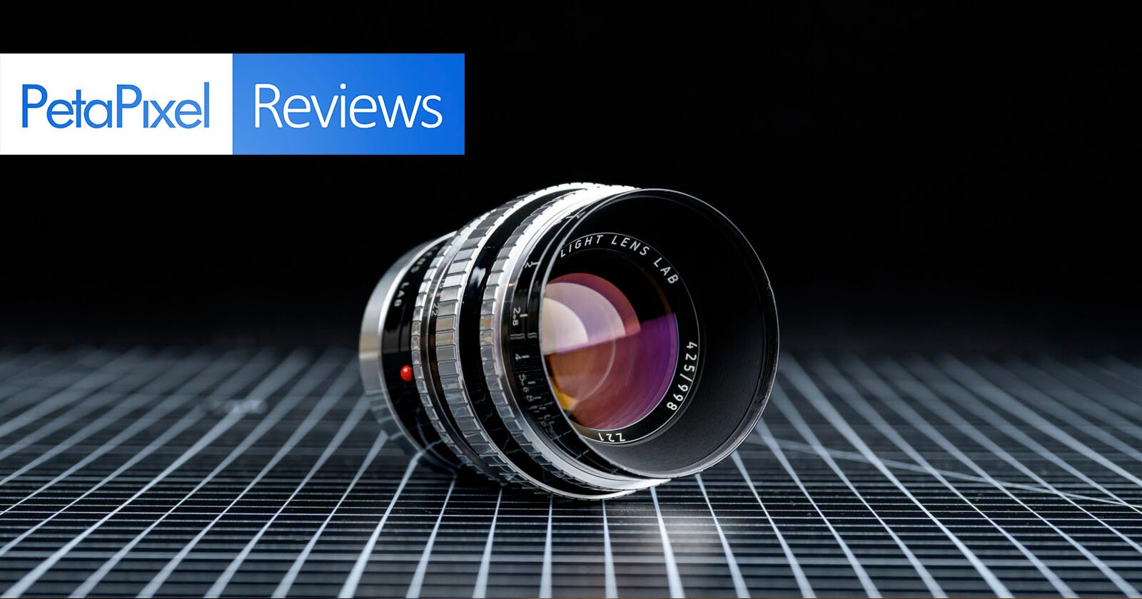 A camera lens rests on a grid-patterned surface against a black background. In the top left corner, the PetaPixel Reviews logo is displayed with blue and white text. A camera lens rests on a grid-patterned surface against a black background. In the top left corner, the PetaPixel Reviews logo is displayed with blue and white text.