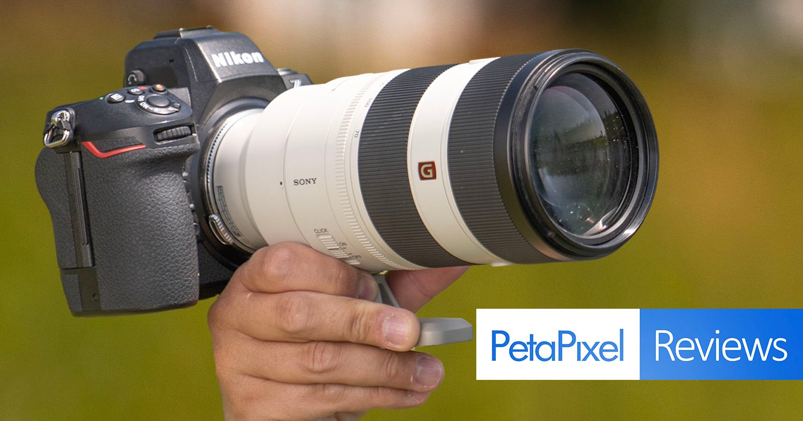 A hand holds a Nikon camera body attached to a large white Sony G Master zoom lens with a blurred outdoor background. The image includes the Petapixel Reviews logo in the lower right corner. A hand holds a Nikon camera body attached to a large white Sony G Master zoom lens with a blurred outdoor background. The image includes the Petapixel Reviews logo in the lower right corner.
