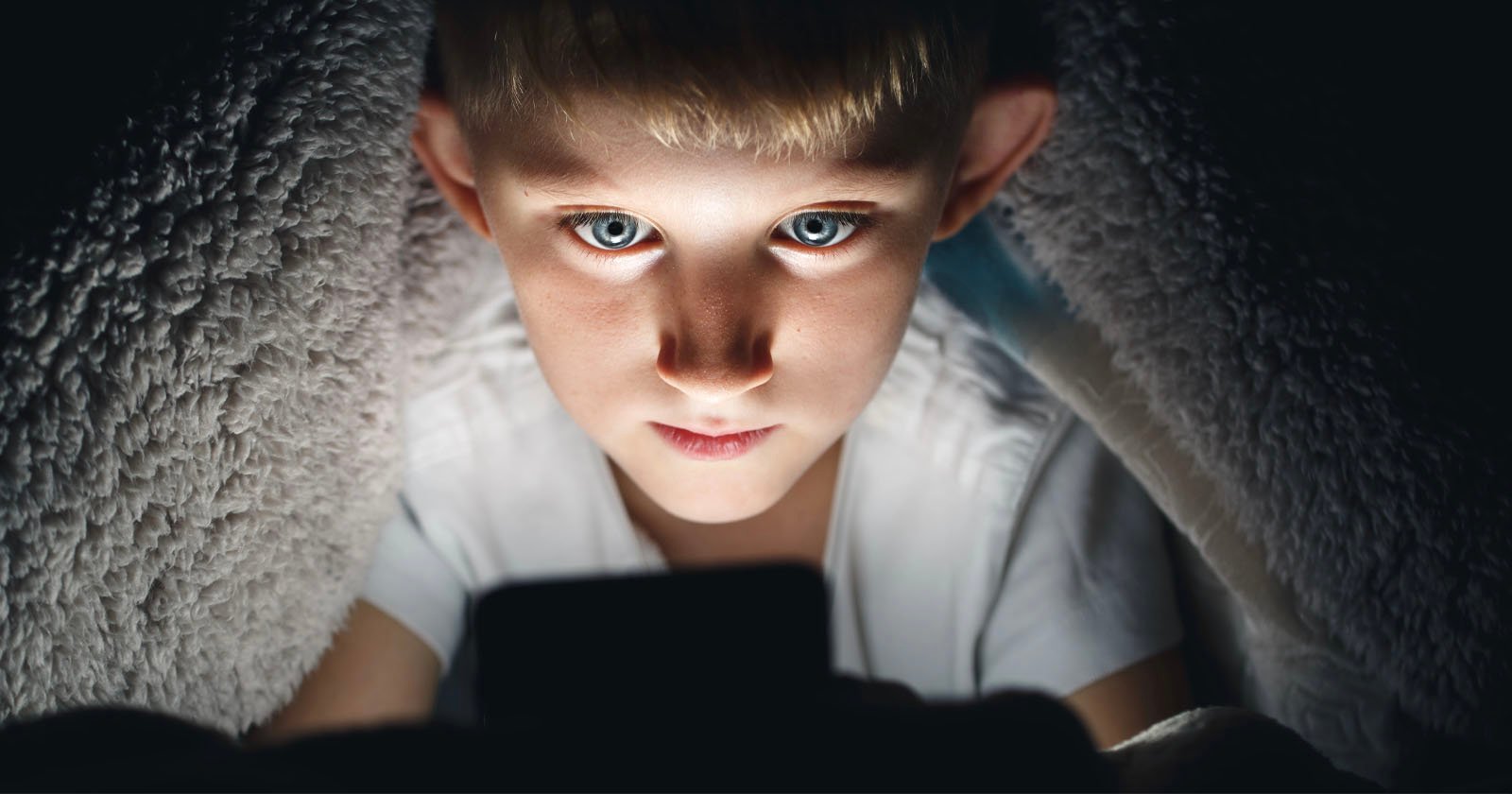 A young boy with blue eyes sits under a blanket, illuminated by the light from a smartphone or tablet he is holding, staring intently at the screen in a dark environment. A young boy with blue eyes sits under a blanket, illuminated by the light from a smartphone or tablet he is holding, staring intently at the screen in a dark environment.
