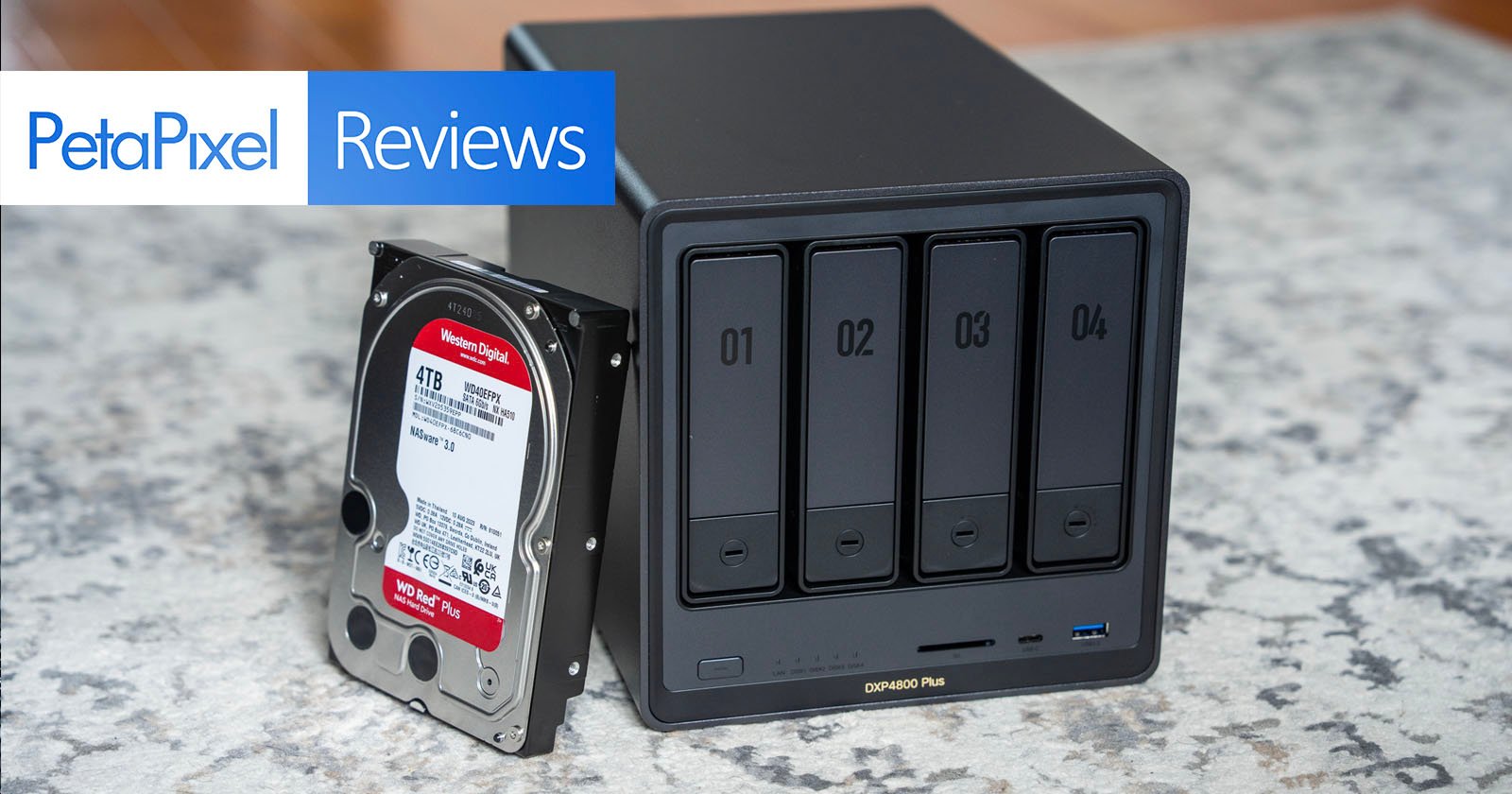 A black four-bay network attached storage device sits on a carpet next to a 4TB hard drive. A “PetaPixel Reviews” banner appears in the upper left corner of the image. A black four-bay network attached storage device sits on a carpet next to a 4TB hard drive. A “PetaPixel Reviews” banner appears in the upper left corner of the image.