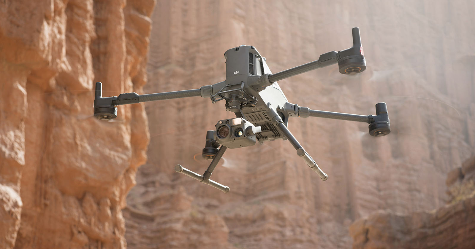 A quadcopter drone equipped with cameras and sensors hovers in front of tall, rugged red rock formations in a canyon, with sunlight streaming through the scene. A quadcopter drone equipped with cameras and sensors hovers in front of tall, rugged red rock formations in a canyon, with sunlight streaming through the scene.