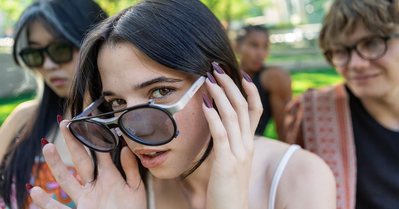 A young woman with long nails adjusts her sunglasses and looks at the camera, with two friends in the background, outdoors on a sunny day in a park. A young woman with long nails adjusts her sunglasses and looks at the camera, with two friends in the background, outdoors on a sunny day in a park.