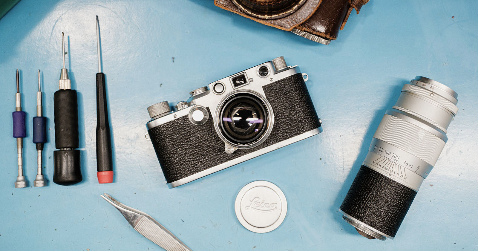 A vintage Leica camera sits on a blue surface surrounded by screwdrivers, tweezers, a camera lens, and a lens cap, suggesting camera repair or maintenance. A vintage Leica camera sits on a blue surface surrounded by screwdrivers, tweezers, a camera lens, and a lens cap, suggesting camera repair or maintenance.