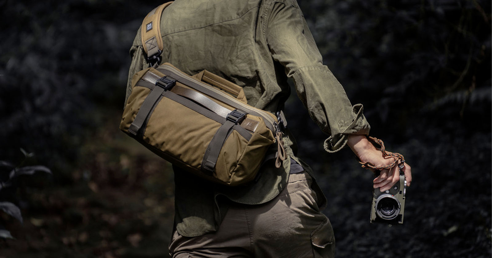 A person wearing olive-green outdoor clothing carries a khaki shoulder bag and holds a camera while walking through a dark, forested area. A person wearing olive-green outdoor clothing carries a khaki shoulder bag and holds a camera while walking through a dark, forested area.