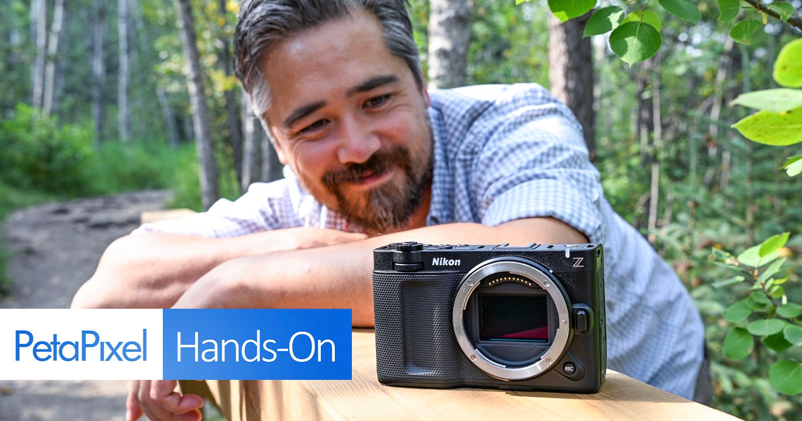 A man with a beard smiles while leaning on a wooden railing in a forest, looking at a Nikon Z camera body placed in front of him. The image includes the text “PetaPixel Hands-On.”. A man with a beard smiles while leaning on a wooden railing in a forest, looking at a Nikon Z camera body placed in front of him. The image includes the text “PetaPixel Hands-On.”.