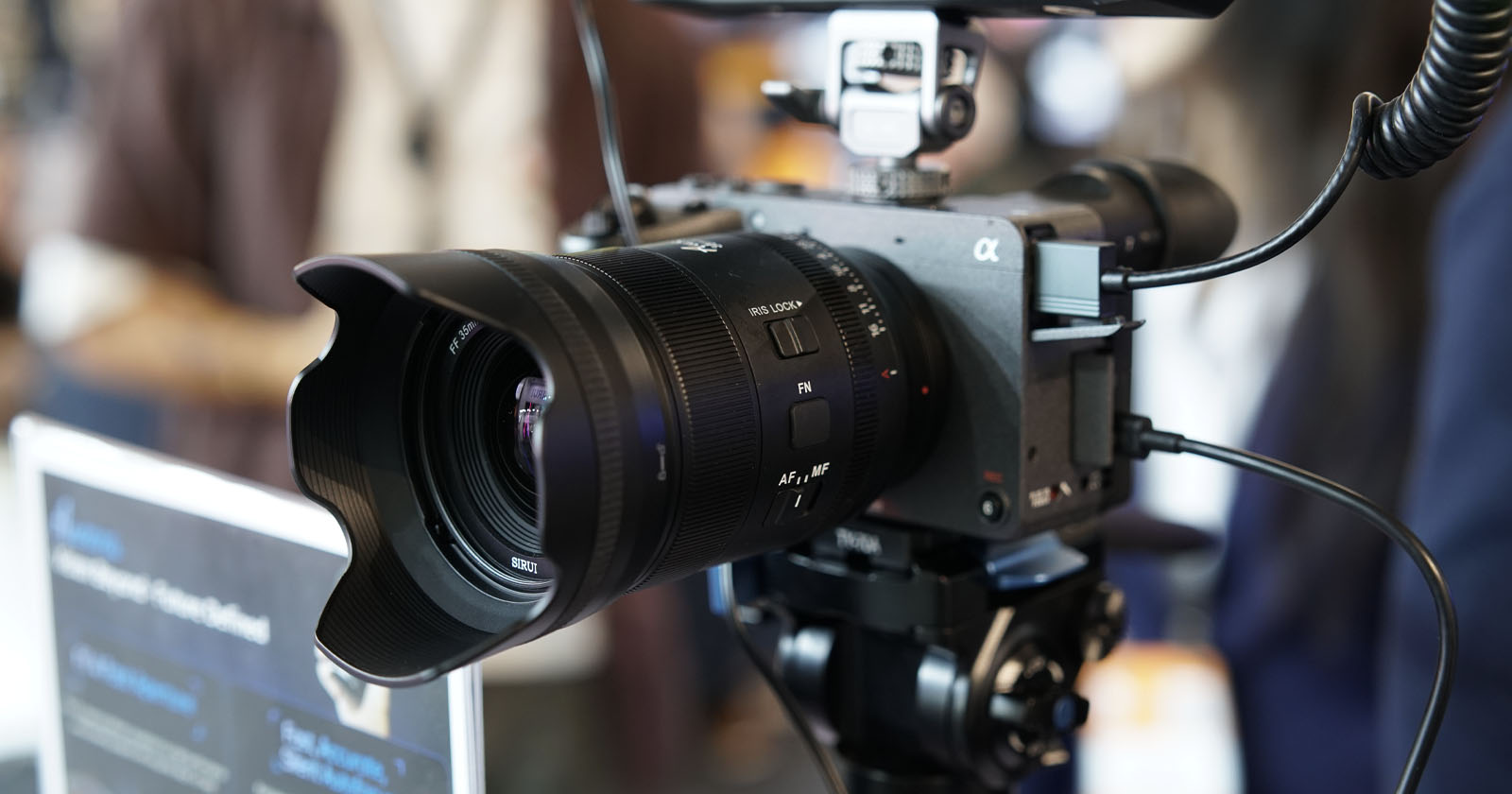Close-up of a professional digital camera with a large lens and lens hood mounted on a tripod, surrounded by cables, set up indoors with a blurred background. Close-up of a professional digital camera with a large lens and lens hood mounted on a tripod, surrounded by cables, set up indoors with a blurred background.