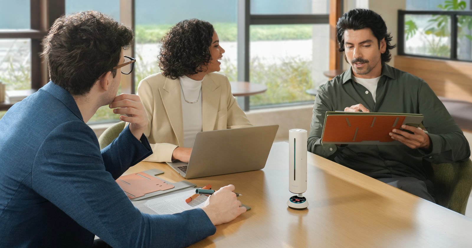 Three people sit around a table in an office, having a discussion. One person writes on a notepad, another listens with a laptop open, and a third observes. A small, white conference microphone sits on the table. Three people sit around a table in an office, having a discussion. One person writes on a notepad, another listens with a laptop open, and a third observes. A small, white conference microphone sits on the table.