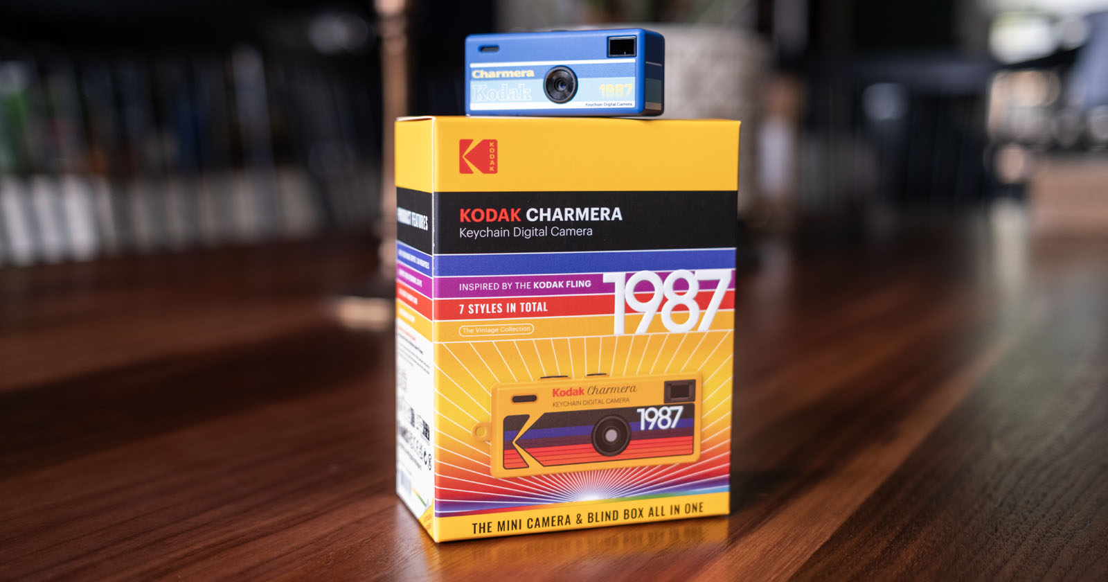 A blue Kodak Charmera keychain digital camera is placed on top of its colorful yellow and red box, which features bold 1987 text and graphics inspired by retro Kodak film packaging. A blue Kodak Charmera keychain digital camera is placed on top of its colorful yellow and red box, which features bold 1987 text and graphics inspired by retro Kodak film packaging.