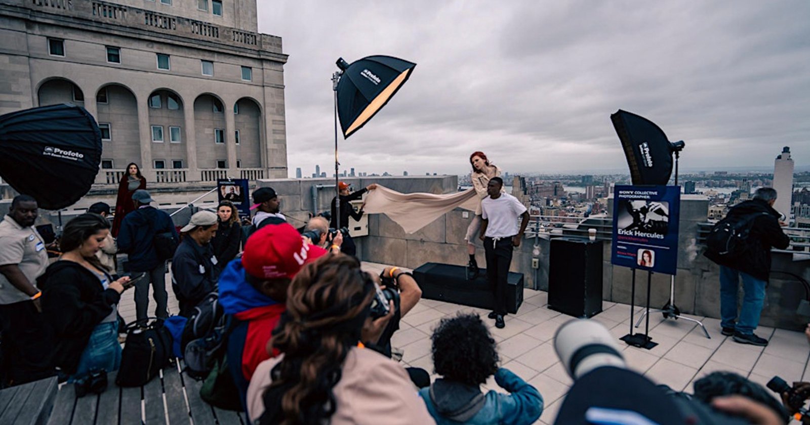 A rooftop photoshoot scene with a crowd watching as a photographer captures two models, one standing and one tossing fabric, against a cityscape. Large lights and camera equipment are set up around them. A rooftop photoshoot scene with a crowd watching as a photographer captures two models, one standing and one tossing fabric, against a cityscape. Large lights and camera equipment are set up around them.