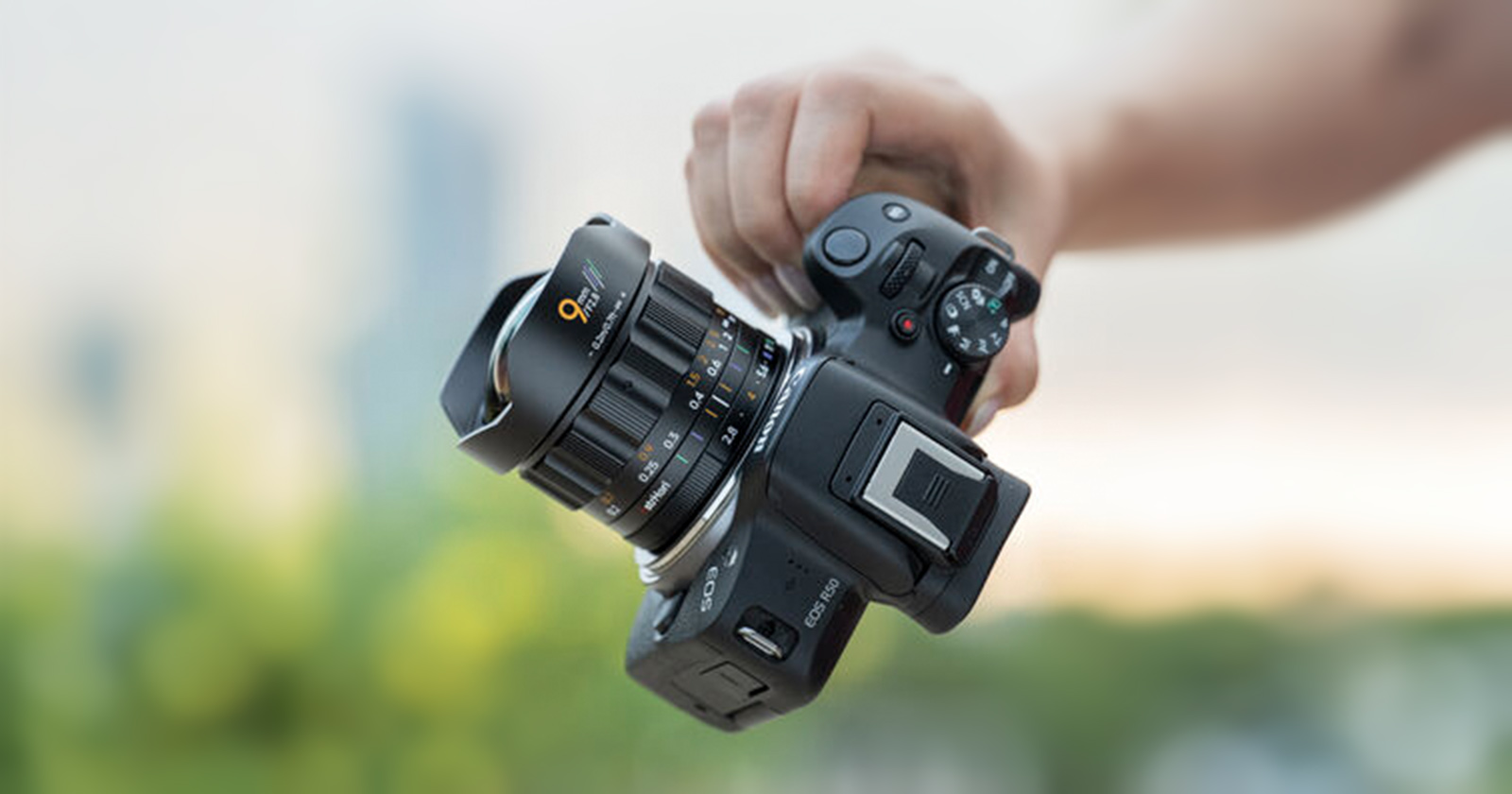 A person holds a black digital camera with a large lens in their right hand; the background is blurred with hints of green and blue. A person holds a black digital camera with a large lens in their right hand; the background is blurred with hints of green and blue.