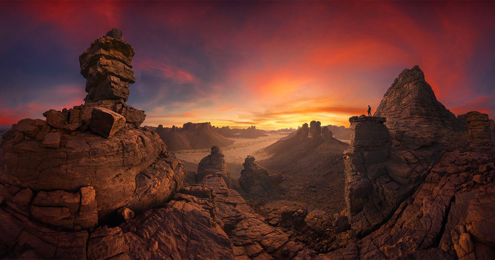 A person stands on a jagged rock formation, overlooking a dramatic, rocky desert landscape at sunset, with vivid orange and purple clouds filling the sky. A person stands on a jagged rock formation, overlooking a dramatic, rocky desert landscape at sunset, with vivid orange and purple clouds filling the sky.
