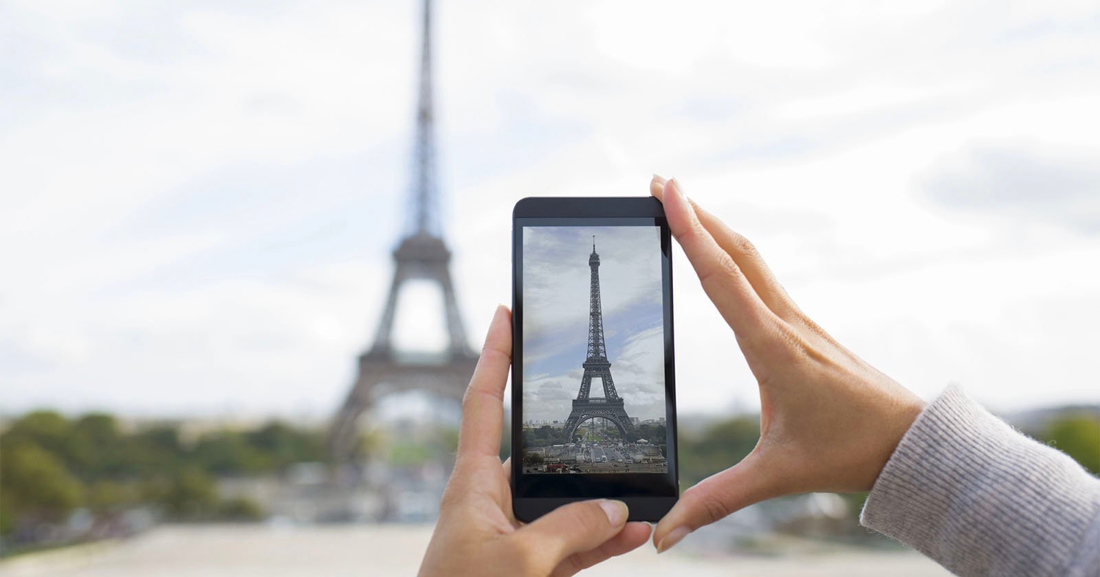 A person's hands hold a smartphone, taking a picture of the Eiffel Tower in Paris. The landmark is blurred in the background but clearly visible on the phone screen. A person's hands hold a smartphone, taking a picture of the Eiffel Tower in Paris. The landmark is blurred in the background but clearly visible on the phone screen.
