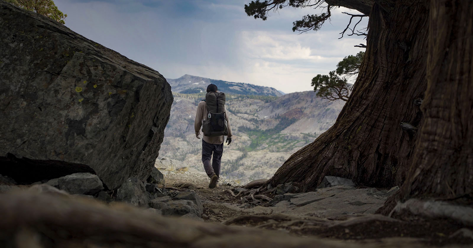 A hiker with a large backpack walks toward a scenic mountain vista, framed by a tree and a large rock, under a partly cloudy sky. A hiker with a large backpack walks toward a scenic mountain vista, framed by a tree and a large rock, under a partly cloudy sky.