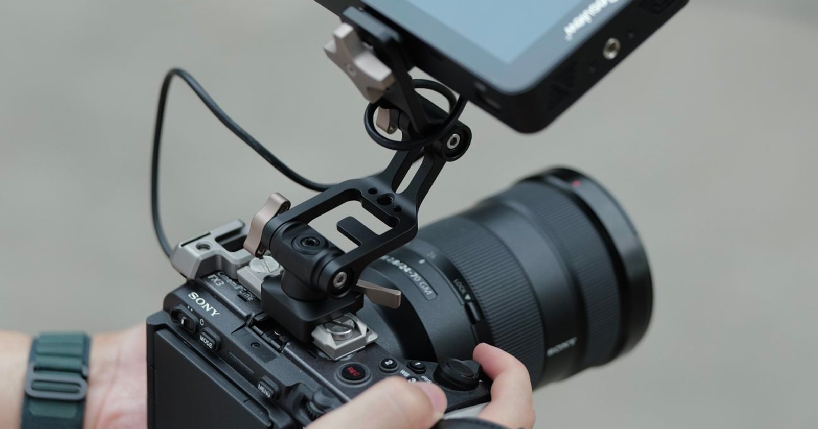 A person holds a Sony camera equipped with a large lens and an external monitor mounted on top, connected by a cable. The camera setup is being adjusted or prepared for use. A person holds a Sony camera equipped with a large lens and an external monitor mounted on top, connected by a cable. The camera setup is being adjusted or prepared for use.