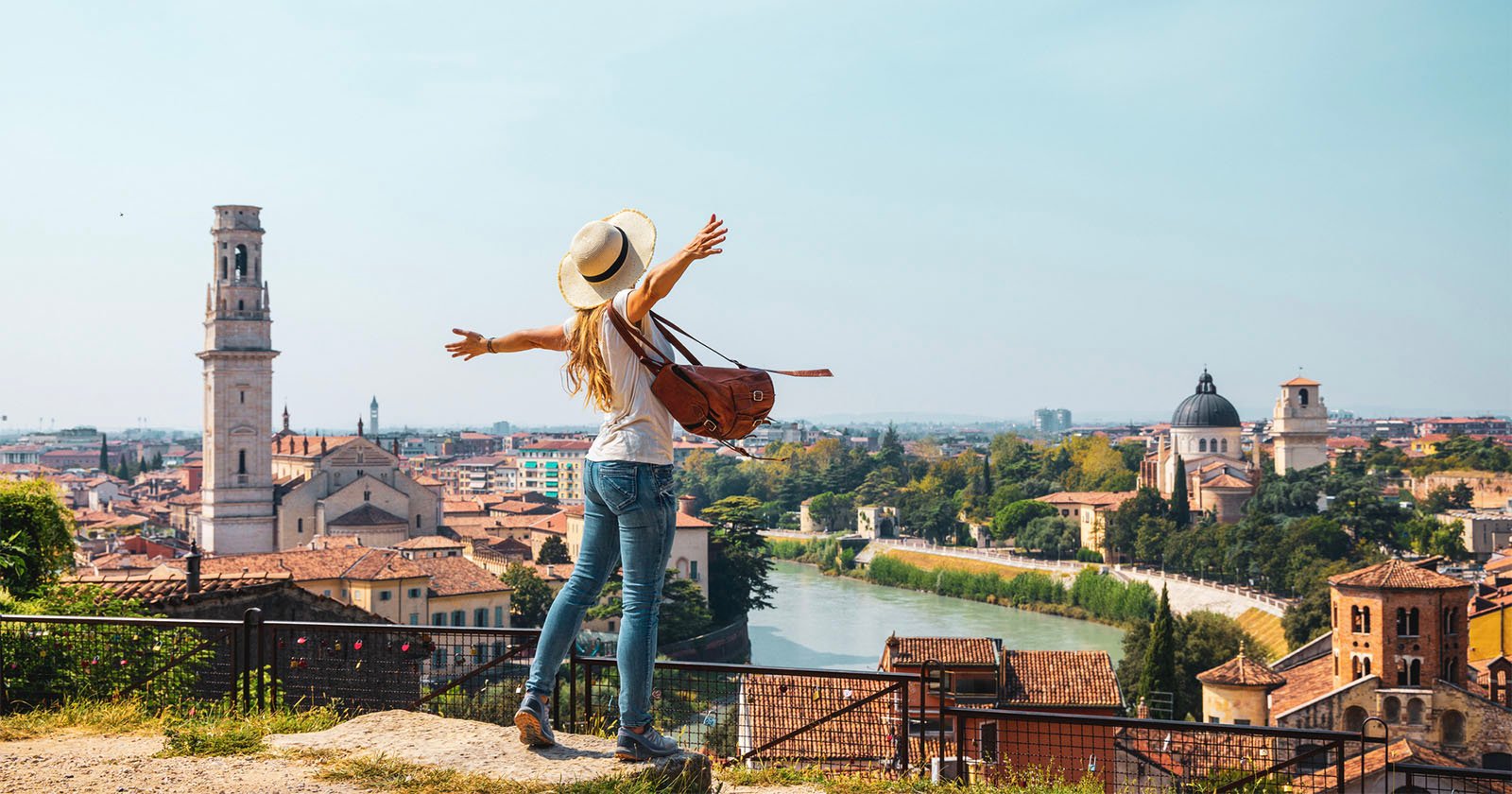 A woman with a hat and backpack stands on a hill with arms outstretched, overlooking a scenic city with historic buildings, a river, and a clear sky. A woman with a hat and backpack stands on a hill with arms outstretched, overlooking a scenic city with historic buildings, a river, and a clear sky.