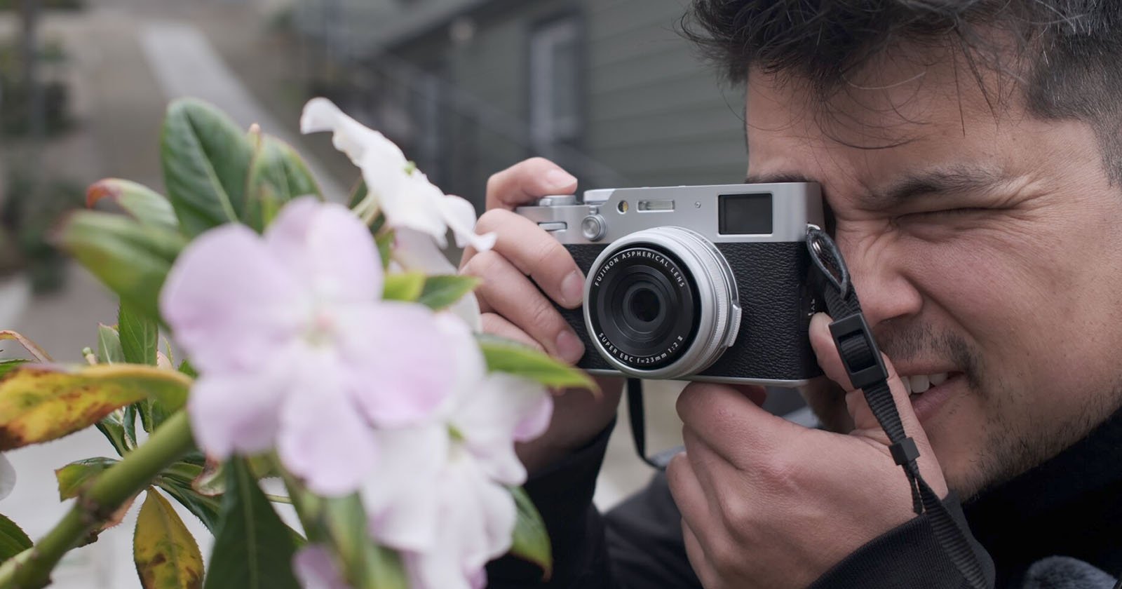 A person holds a camera up to their face, focusing and preparing to take a photo of light pink flowers on a leafy green plant outdoors. A person holds a camera up to their face, focusing and preparing to take a photo of light pink flowers on a leafy green plant outdoors.