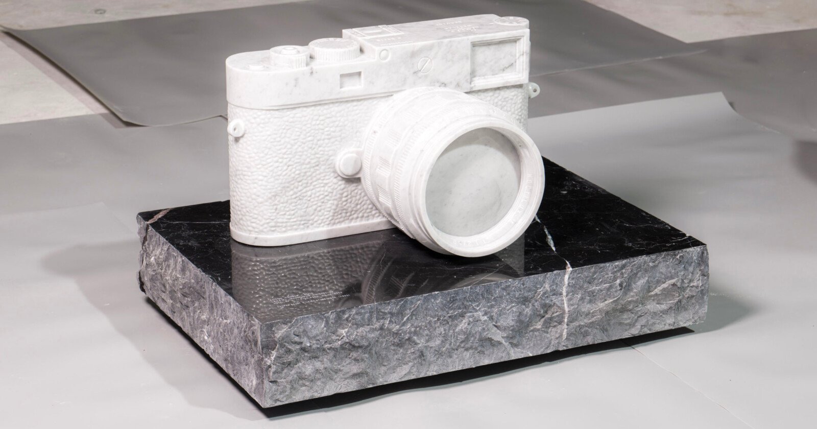 A realistic camera sculpture made of white marble sits on a rough, rectangular black stone base, displayed on a neutral surface background. A realistic camera sculpture made of white marble sits on a rough, rectangular black stone base, displayed on a neutral surface background.