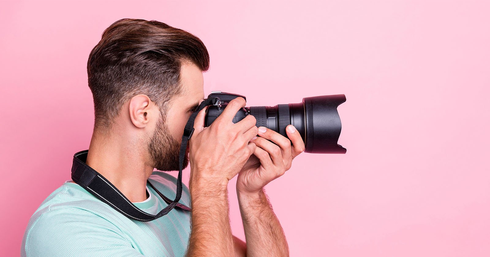 A man with short brown hair and a beard is holding a professional camera up to his face, taking a photo against a pink background. He is wearing a mint green t-shirt and has a camera strap around his neck. A man with short brown hair and a beard is holding a professional camera up to his face, taking a photo against a pink background. He is wearing a mint green t-shirt and has a camera strap around his neck.